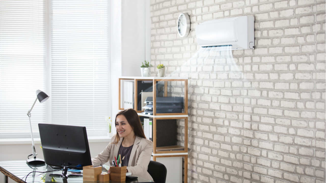 Ar-condicionado split branco ligado na parede de um escritório, enquanto mulher branca trabalha no computador em mesa de escritório